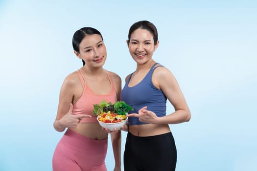 Two young sporty Asian women in sportswear holding salad bowl fill with fruit and vegetable. Natural youthful and fit body lifestyle people with balance nutrition on isolated background. Vigorous