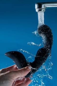 A woman washes a black prostate stimulator under running water on a blue background. Sex toy hygiene concept