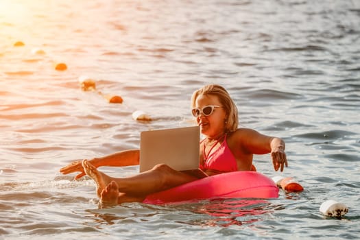 Woman works on laptop in sea. Freelancer, young blond woman in sunglases floating on an inflatable big pink donut with a laptop in the sea at sunset. Freelance, travel and holidays concept