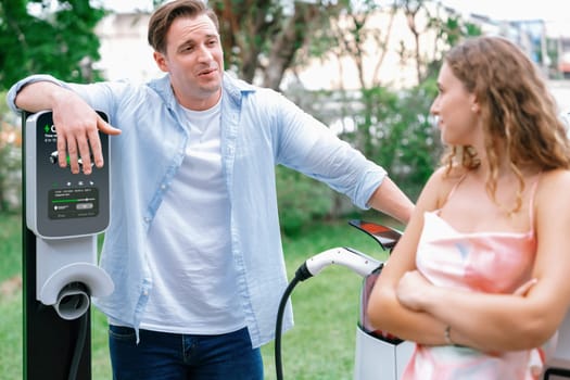 Happy and lovely couple with eco-friendly conscious recharging electric vehicle from EV charging station. EV car technology utilized as alternative transportation for future sustainability. Synchronos