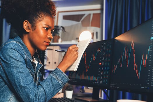 Concentrated young African woman trader writing analysis, looking on screen with stock market chart at high profit. Concept of exchange investment online in trading application on pc. Tastemaker.