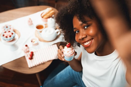 Beautiful young African blogger presenting piece of cupcake in concept special cuisine with selfie on smartphone. Content creating of social media with favorite sweets bakery dish. Tastemaker.