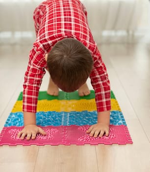 Sport and health concept. A little boy 4 years old in red checkered pajamas is having fun on a multi-colored massage orthopedic mat with spikes in a home interior. Close-up. Soft focus.