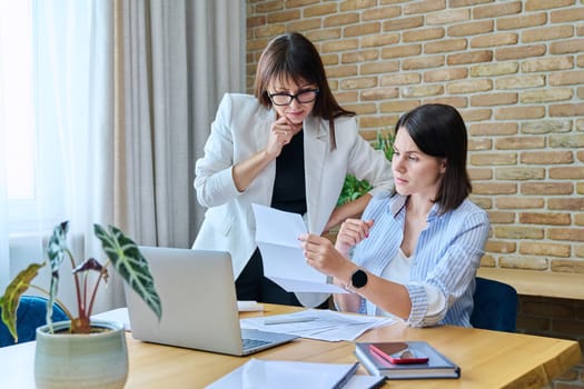 Two serious business women talking working in modern office, young female reading contract document, communicating colleague. Business, ceo, work, law, finance, mentoring, consulting, teamwork, job concept