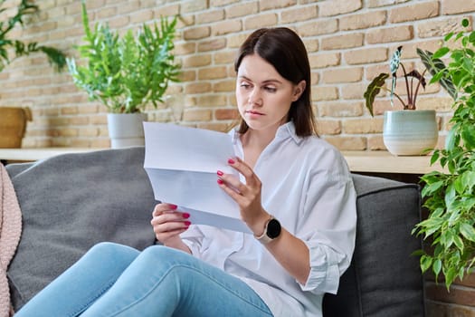 Serious young woman reading official paper letter while sitting on sofa in living room at home. Notification from bank, work, social tax services, invoice, insurance company, about rental property
