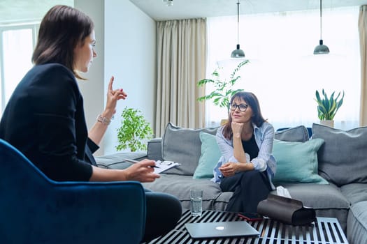 Female psychologist is having therapy session with middle-aged woman patient sitting on sofa in office. Psychology, psychotherapy, therapy, counseling, treatment, mental health concept