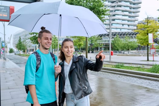 Teenagers guy and girl students with backpacks at bus stop under an umbrella waiting for city bus. Public transport, adolescence, youth, city life, lifestyle, urban style.