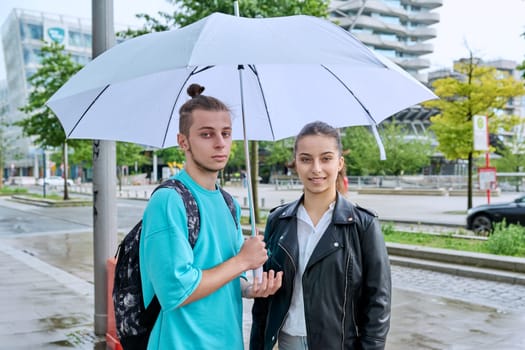 Outdoor portrait of teenagers students friends smiling guy and girl looking at camera under an umbrella on street of modern city. College, high school, urban lifestyle, adolescence, youth concept