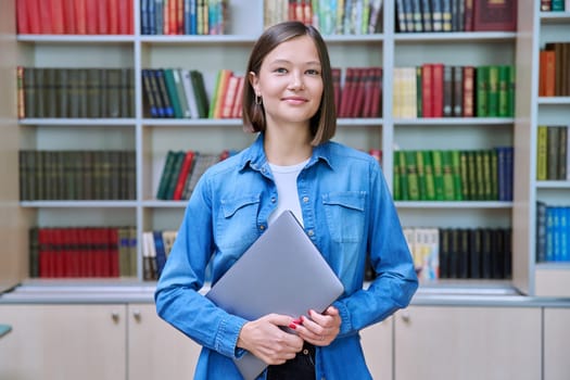 Portrait of smiling young female university student looking at camera, standing posing with laptop in library classroom of educational building. Knowledge, higher education, youth concept