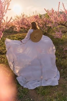 Woman blooming peach orchard. Against the backdrop of a picturesque peach orchard, a woman in a long white dress and hat enjoys a peaceful walk in the park, surrounded by the beauty of nature