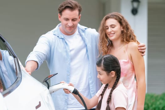 Happy little young girl learn about eco-friendly and energy sustainability as she help her family recharge electric vehicle from home EV charging station. EV car and modern family concept. Synchronos