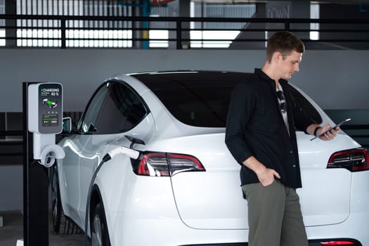 Young man travel with EV electric car to shopping center parking lot charging in downtown city showing urban sustainability lifestyle by green clean rechargeable energy of electric vehicle innards