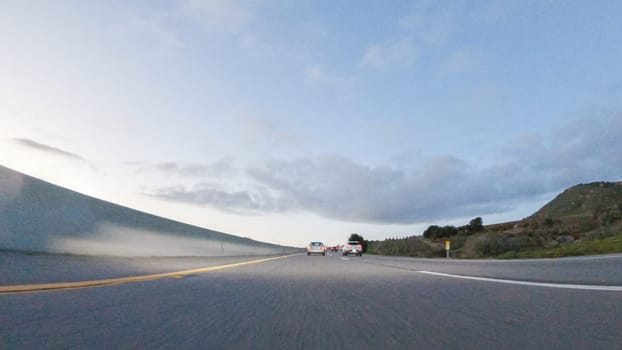 As the sun sets, driving on HWY 101 near Pismo Beach, California becomes a captivating experience with the sky painted in shades of orange and pink, casting a warm glow over the coastal landscape.