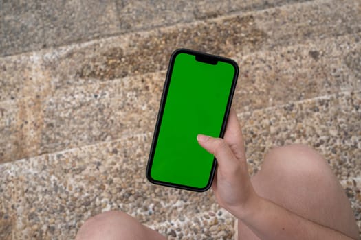 Top view of anonymous young person looking at chromakey screen of mobile phone and browsing information while sitting outside on marble steps in daylight