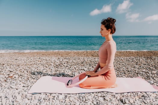 Middle aged well looking woman with black hair doing Pilates with the ring on the yoga mat near the sea on the pebble beach. Female fitness yoga concept. Healthy lifestyle, harmony and meditation.