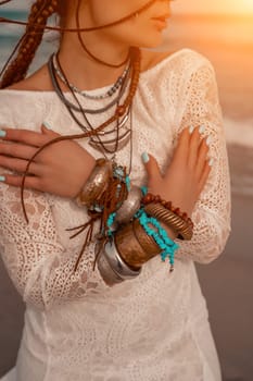 Model in boho style in a white long dress and silver jewelry on the beach. Her hair is braided, and there are many bracelets on her arms