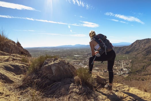 Beautiful mountain standing young woman with backpack on top at sunset in summer. Landscape with a sporty girl, hills, blue sky with sun rays. Travel and tourism. High quality photo