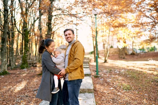 Smiling dad stands next to mom holding little girl in her arms in park. High quality photo