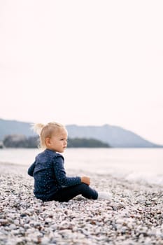 Little girl sits on a pebble beach and looks at the sea. Side view. High quality photo