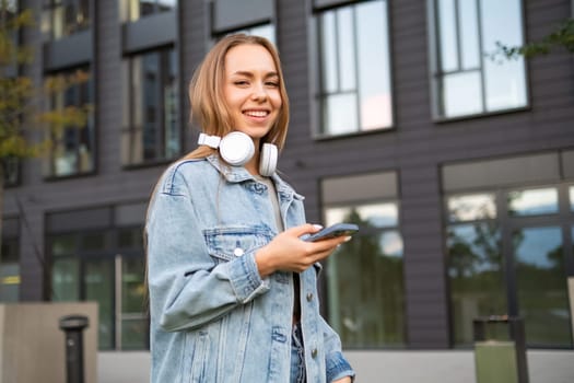 A grinning young woman, with her phone in hand and headphones stylishly draped around her neck, texting to her friend using smartphone