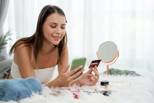 Young woman lying on the bed using online payment app and digital wallet on smartphone to pay with credit card. E commerce shopping and modern purchasing via mobile internet. Unveiling