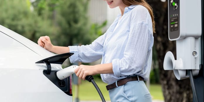 Young woman recharge EV electric vehicle's battery from EV charging station in outdoor green city park scenic. Eco friendly urban transport and commute with eco friendly EV car travel. Panorama Exalt