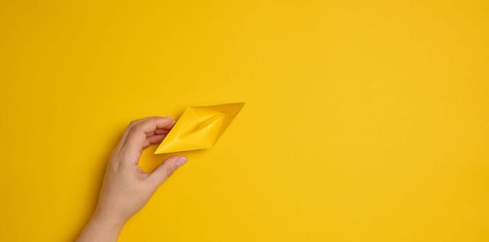 Female hand hold a blue paper boat on a yellow background. Mentoring and support concept, top view
