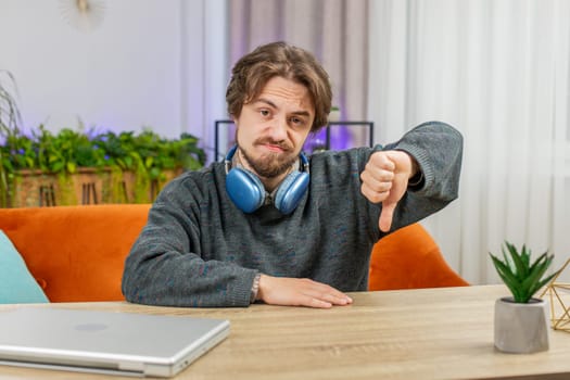 Dislike. Upset unhappy Caucasian man showing thumbs down sign fail gesture, expressing discontent, disapproval dissatisfied bad work at home office indoors. Displeased young guy in room sits at table