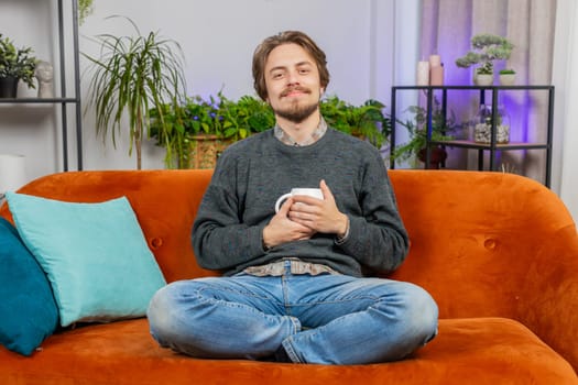 Attractive smiling bearded man drinking a cup of warm coffee or herbal tea sitting at home apartment living room sofa in the morning. Caucasian young adult guy enjoying comfortable relaxing alone