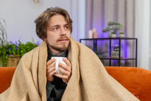 Sick ill man wrapped in plaid sit shivering from cold drinking hot coffee or herbal tea in unheated home apartment without heating due debt. Unhealthy Caucasian guy try to warming up sitting on sofa