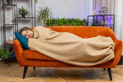 Tired young man lying down in bed taking a rest at home apartment room. Carefree Caucasian guy napping, falling asleep on comfortable orange sofa with pillows. Closed his eyes enjoy daytime nap alone