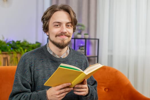 Caucasian young man reading interesting book, turning pages smiling enjoying literature, taking a rest on comfortable couch. Portrait of peaceful cheerful guy relaxing at home apartment living room