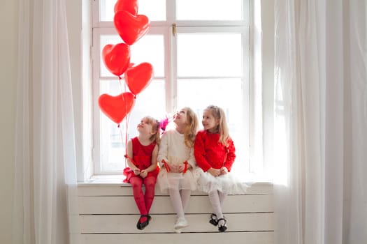 Three little girls sitting on a window with red heart balloons