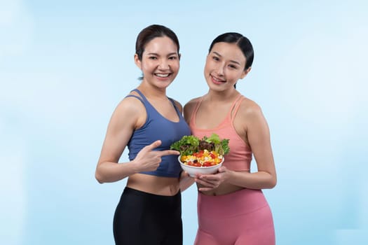 Two young sporty Asian women in sportswear holding salad bowl fill with fruit and vegetable. Natural youthful and fit body lifestyle people with balance nutrition on isolated background. Vigorous