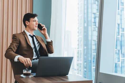 Closeup of handsome businessman making phone call with manager while sitting near window with skyscraper view. Executive manager talking working by using phone and laptop. Look aside. Ornamented.