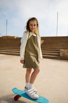 Full length portrait of cute child girl dressed in stylish khaki dress and beige hoodie, standing on her skateboard on one leg, smiling looking at the camera, enjoying skateboarding on the playground