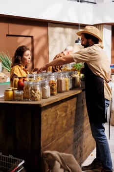 Client shopping for nutritious organic pantry staples in zero waste supermarket, being assisted with information by trader. Vendor showing green living woman locally sourced products