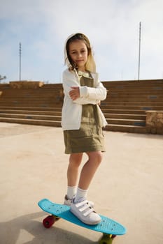 Caucasian adorable confident kid girl, 6 years old, dressed in stylish khaki dress and creamy hoody, looking at camera, standing with arms folded on her skateboard on one leg on the urban playground