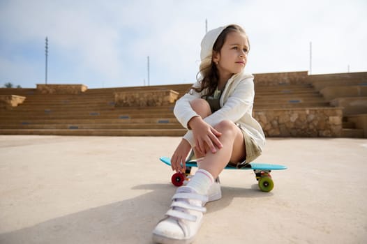Adorable Caucasian little kid girl sitting on her skateboard, dreamily looking away. People. Childhood. Leisure activity.