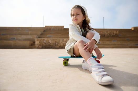 Beautiful Caucasian elementary age child girl in khaki dress and beige hoodie, dreamily looking away, sitting on a blue skateboard with multicolored wheels, on urban skatepark. Kids and extreme sport