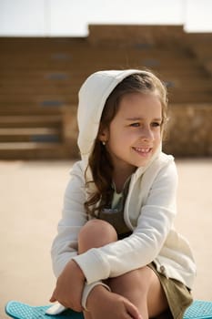 Close-up portrait of a Caucasian elementary age stylish kid girl sitting on her blue skateboard, smiling dreamily looking away, dressed in beige hoodie. Childhood, Leisure activity. Active lifestyle