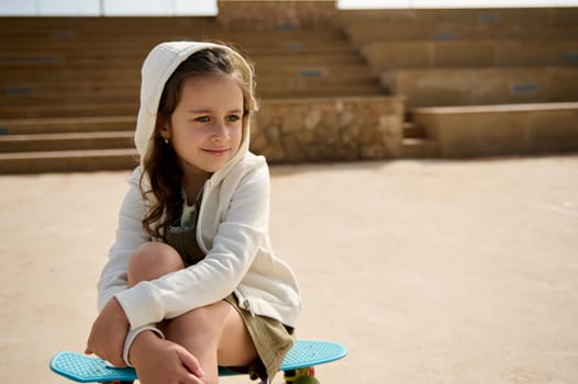 Beautiful elementary age Caucasian child, adorable little girl with hood on her head, smiles cutely and dreamily looking away, sitting on skateboard outdoor on urban skatepark against steps background