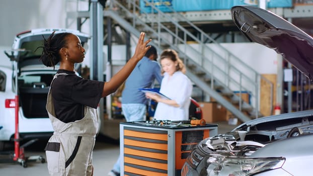 Licensed engineer in auto repair shop using augmented reality hologram to visualize car components in order to fix them. African american woman using AR technology while working on damaged vehicle