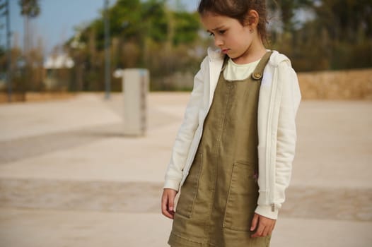 Portrait of a little child girl in khaki dress and beige hoodie, looking down, expressing sadness standing on urban background. People. Lifestyle. Emotional portrait