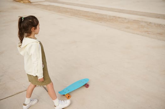 Little child girl in beige hoodie and khaki dress, skateboarding on the urban skatepark or playground. View from above. Happy active childhood. Extreme sport. Leisure activity