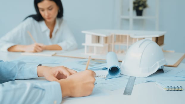 Professional architect engineer drawing blueprint and working together with young beautiful caucasian coworker on meeting table with safety helmet, house model, blueprint scatter around. Immaculate.