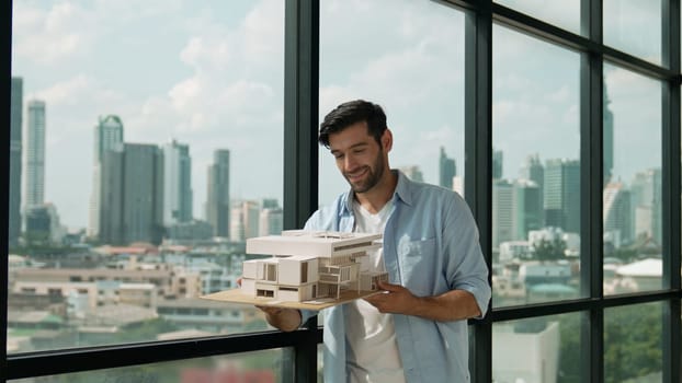 Skilled smiling caucasian engineer turns house model left and right to check mistake point. Professional manager inspect at architectural model while standing near window. Design concept. Tracery