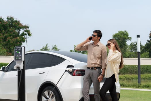 Young couple recharge her EV electric vehicle at green city park parking lot while talking on phone. Sustainable urban lifestyle for environment friendly EV car with battery charging station.Expedient