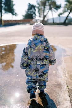 Little girl in rubber boots walks through a puddle, looking down at her feet. Back view. High quality photo