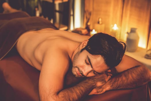 Caucasian man customer enjoying relaxing anti-stress spa massage and pampering with beauty skin recreation leisure in warm candle lighting ambient salon spa at luxury resort or hotel. Quiescent